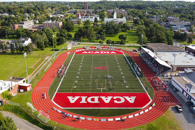 A drone photo of a football game on a sunny day, with people in the stadium stands and the campus in the background.